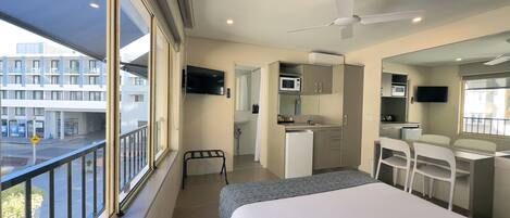 A hotel room with a bed, a desk, a chair, a small kitchenette, a TV, and a balcony with a view of buildings.