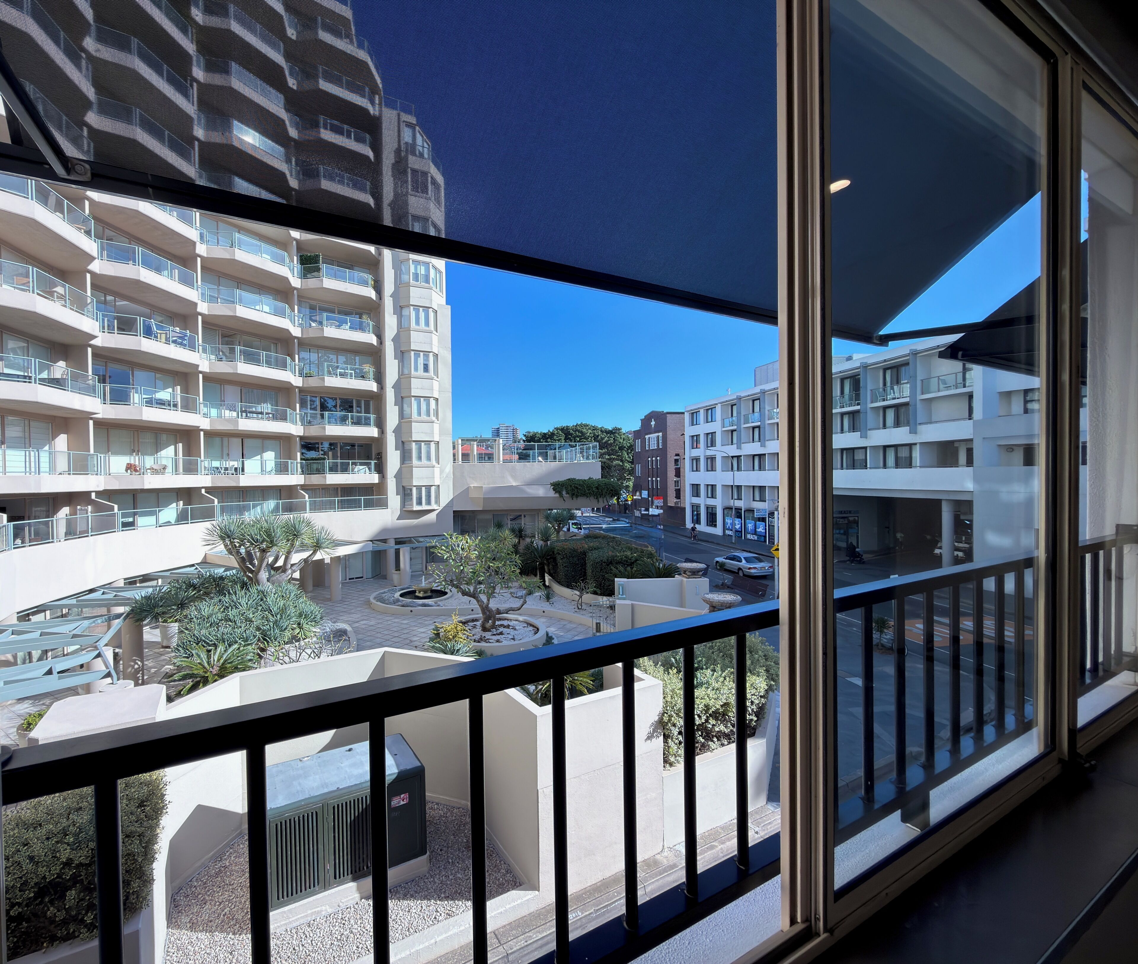 A multi-story residential building with balconies, a courtyard with plants, and a clear blue sky.
