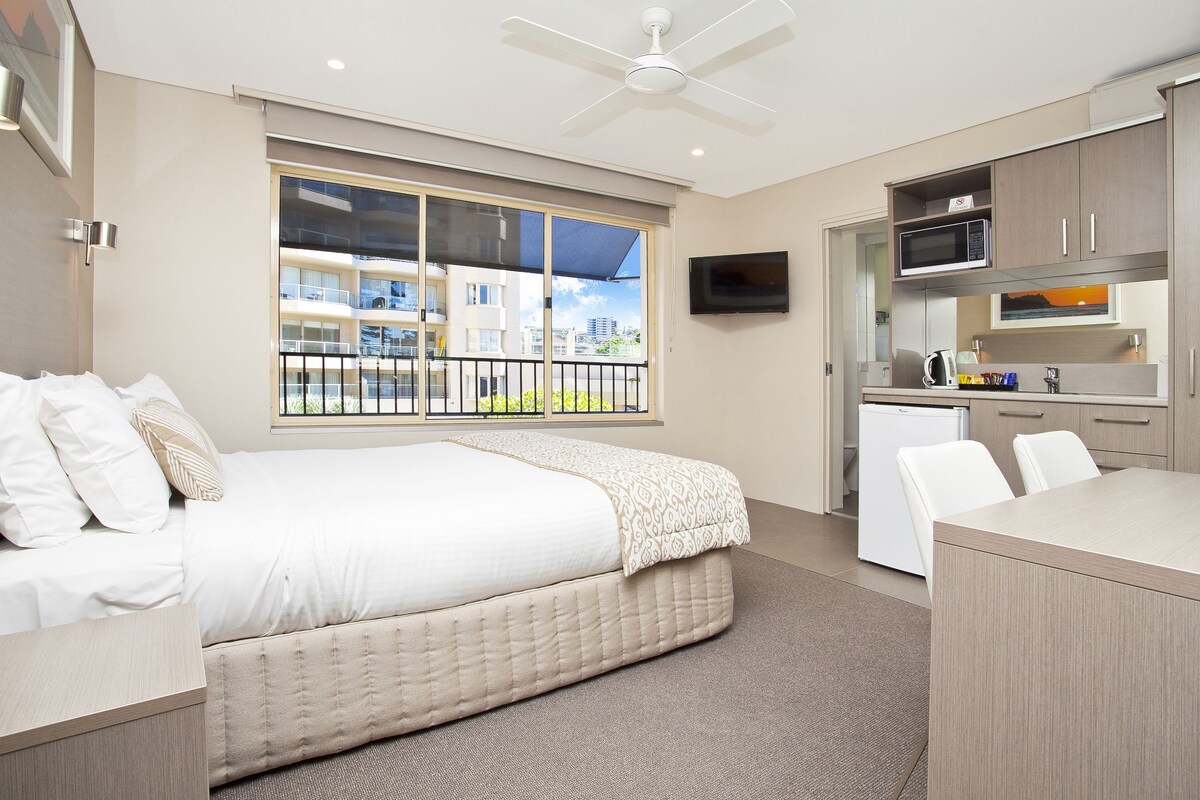 A modern hotel room with a bed, a desk, a kitchenette, and a balcony with a city view.