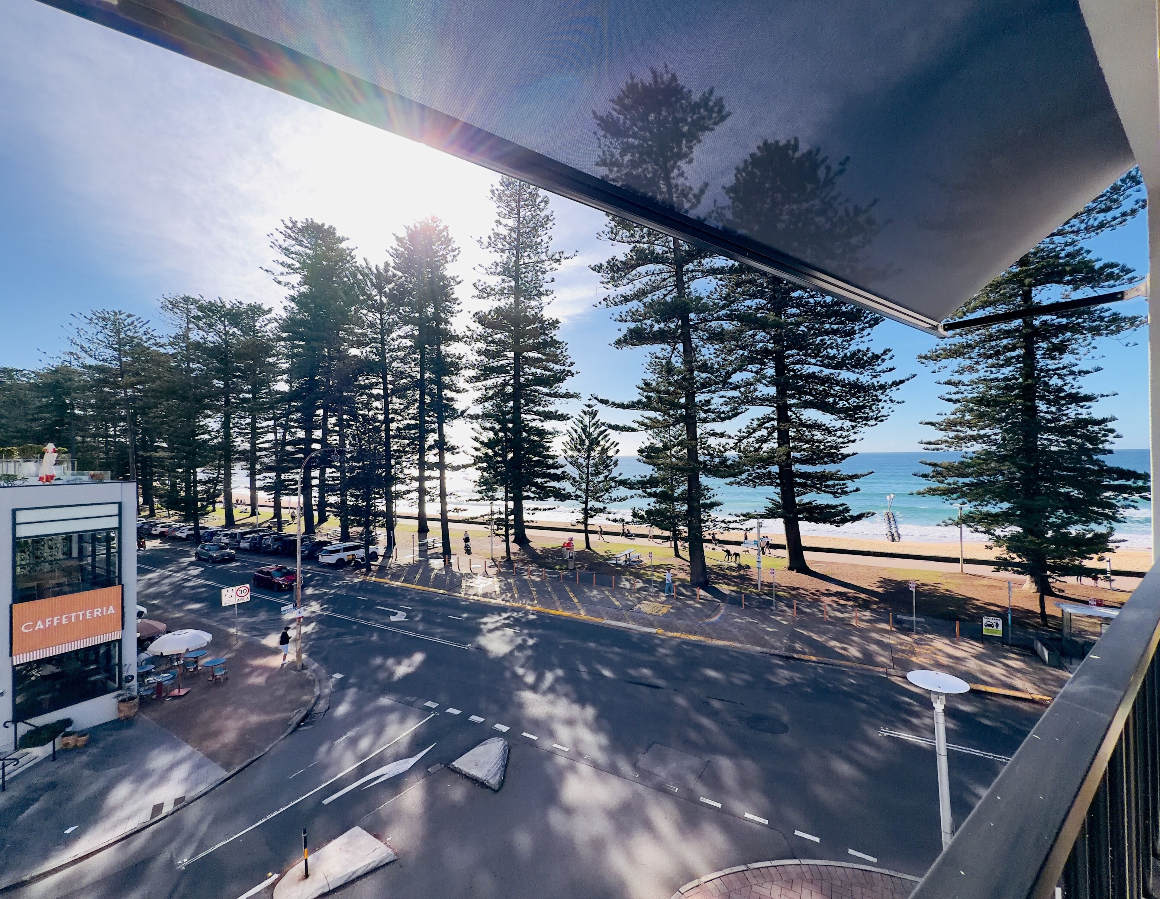 A coastal road with tall trees, a beach in the background, and a building labeled 'CAFFETTERIA' on the left.