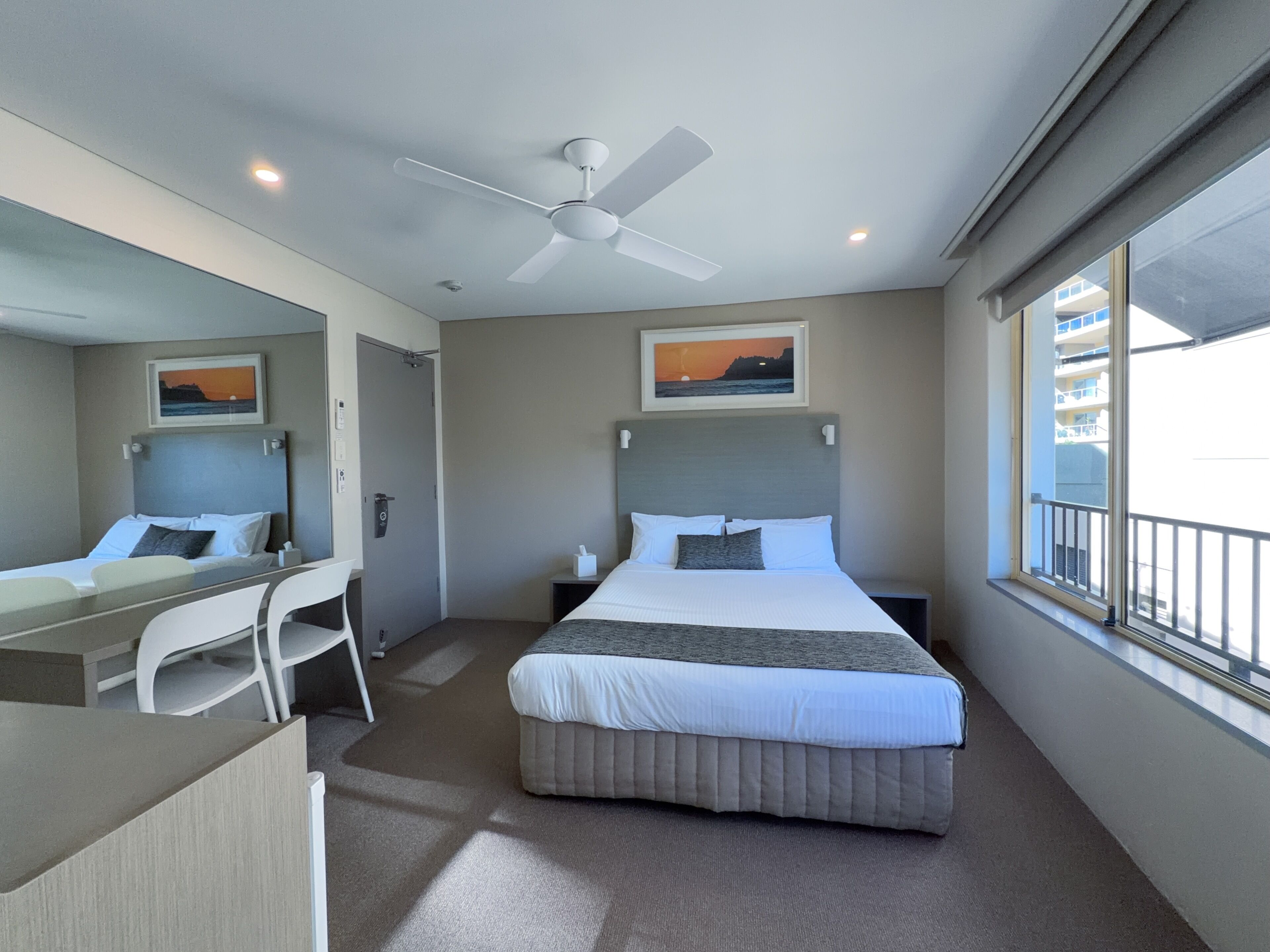 A hotel room with a bed, a desk with a chair, a ceiling fan, a window with a view, and artwork on the walls.