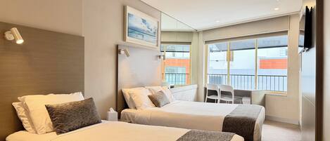 Et hotelværelse med to senge, en ventilator, et skrivebord med stole, et fjernsyn og et stort vindue med udsigt.