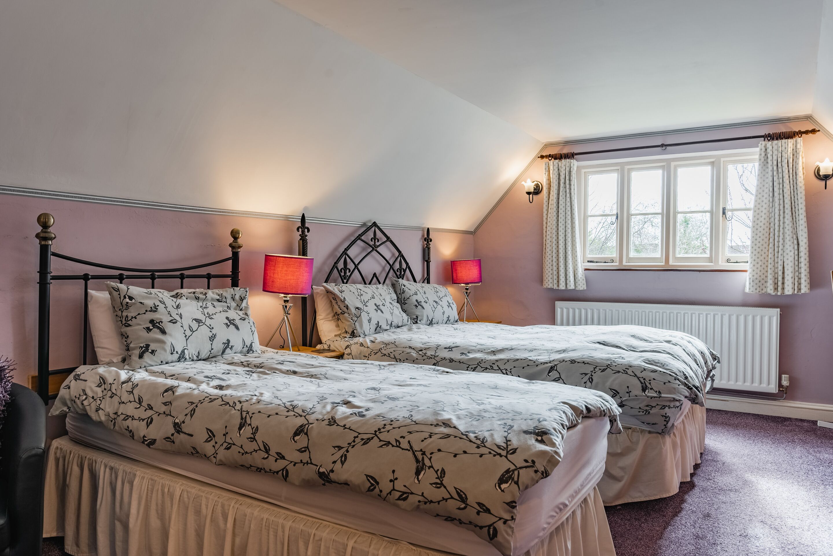 A bedroom with two beds, each with a floral-patterned bedspread and matching pillows. There are two bedside lamps with red shades, and a window with curtains.