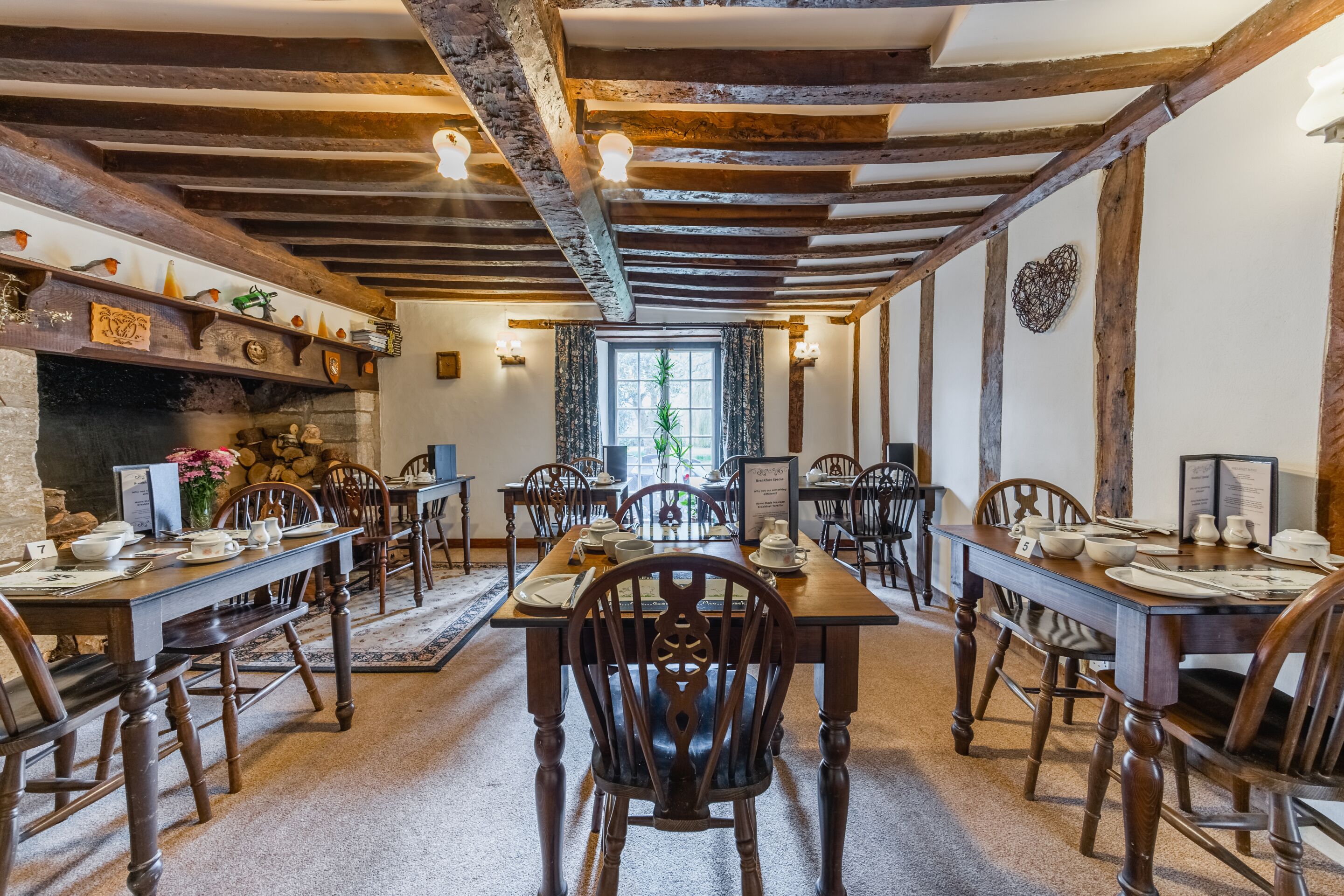 A traditional dining hall with wooden tables and chairs, stone fireplace, and exposed wooden beams.