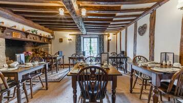 A traditional dining hall with wooden tables and chairs, stone fireplace, and exposed wooden beams.