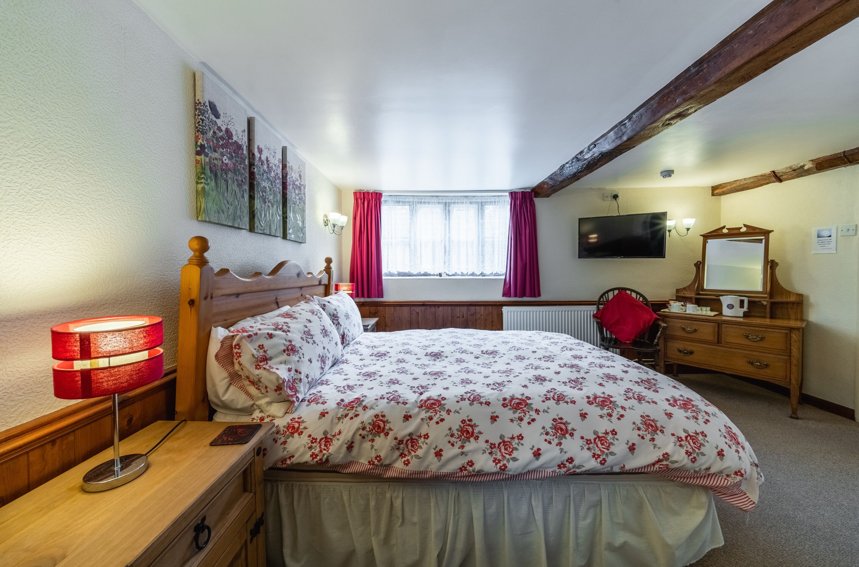 A bedroom with a wooden bed, a nightstand with a lamp, a dresser, a television, and a window with curtains.