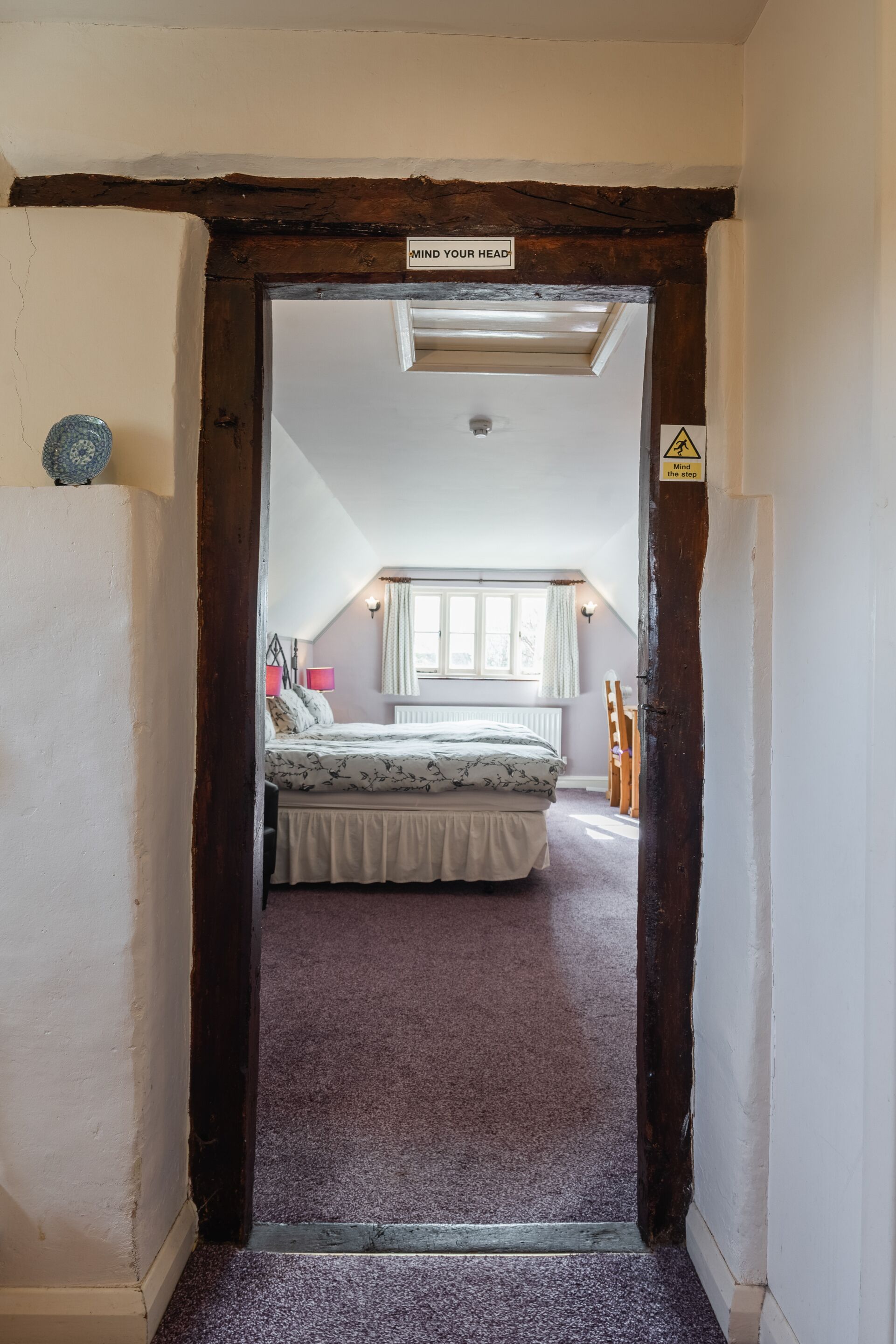 A narrow hallway leading to a bedroom with a bed, a window, and a "Mind Your Head" sign.