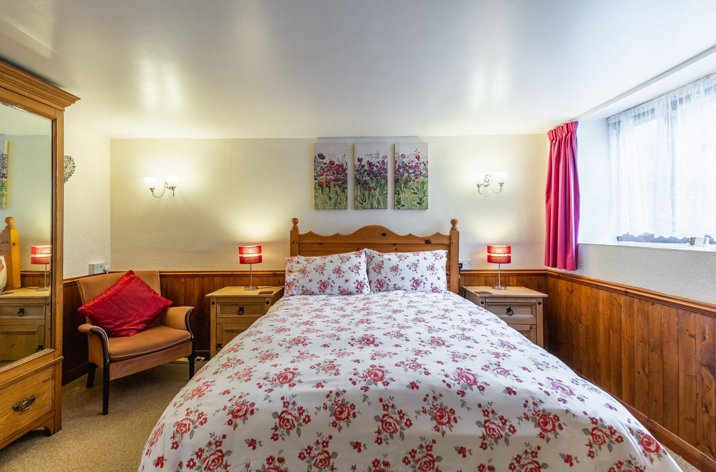 A bedroom with a wooden headboard, floral bedding, two bedside lamps, a chair with a red pillow, and a window with curtains.