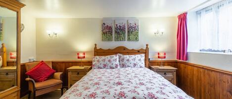 A bedroom with a wooden headboard, floral bedding, two bedside lamps, a chair with a red pillow, and a window with curtains.