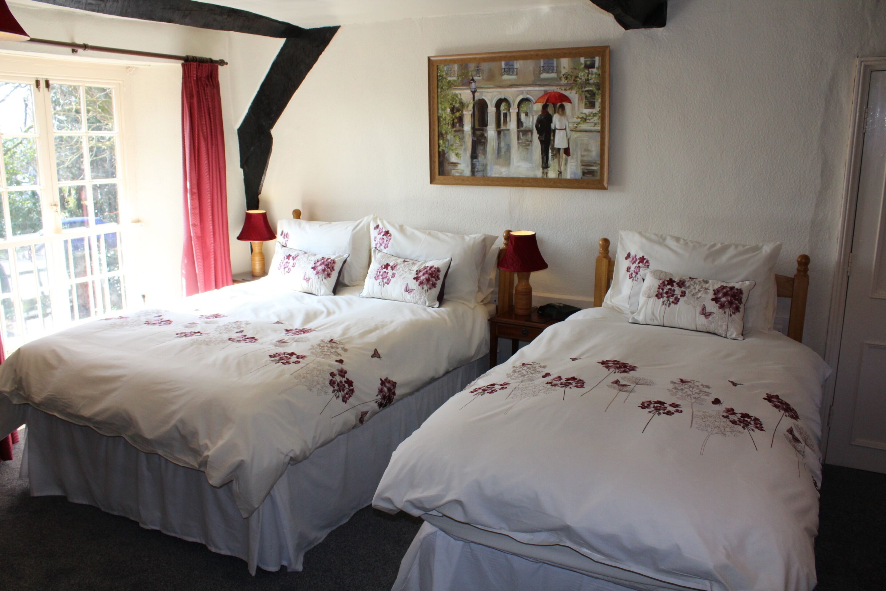 Habitación doble con dos camas, cada una con ropa de cama blanca y motivos florales, un cuadro en la pared y una ventana con cortinas rojas.