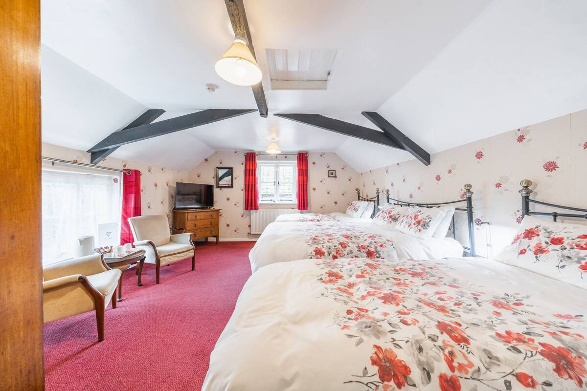 Ein Schlafzimmer mit schrägen Wänden, einem großen Bett mit floraler Bettwäsche, einem Schreibtisch mit Stuhl und einem Fenster mit roten Vorhängen.