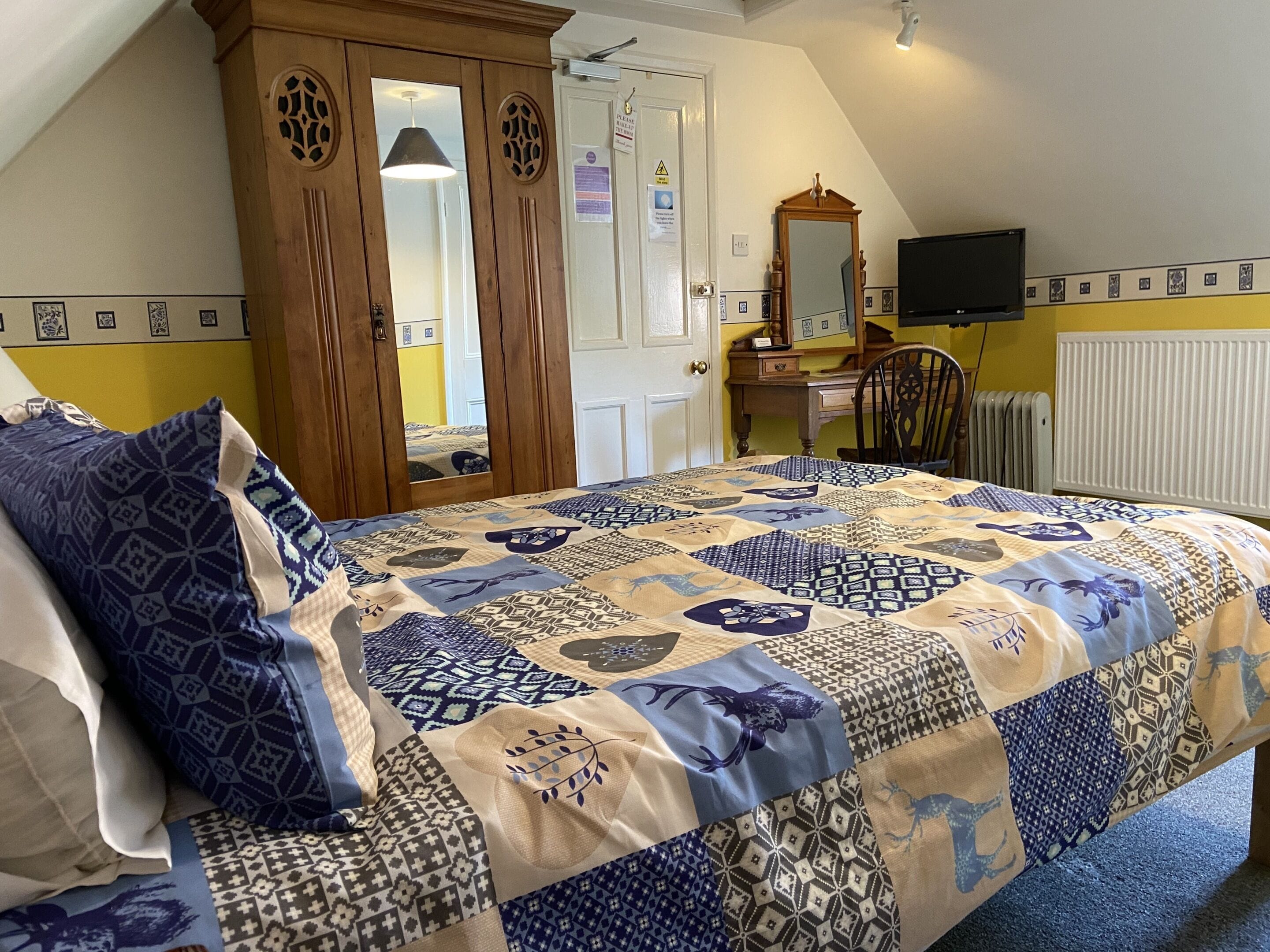 A bedroom with a large bed, a wooden wardrobe, a desk with a chair, a television, and a radiator.