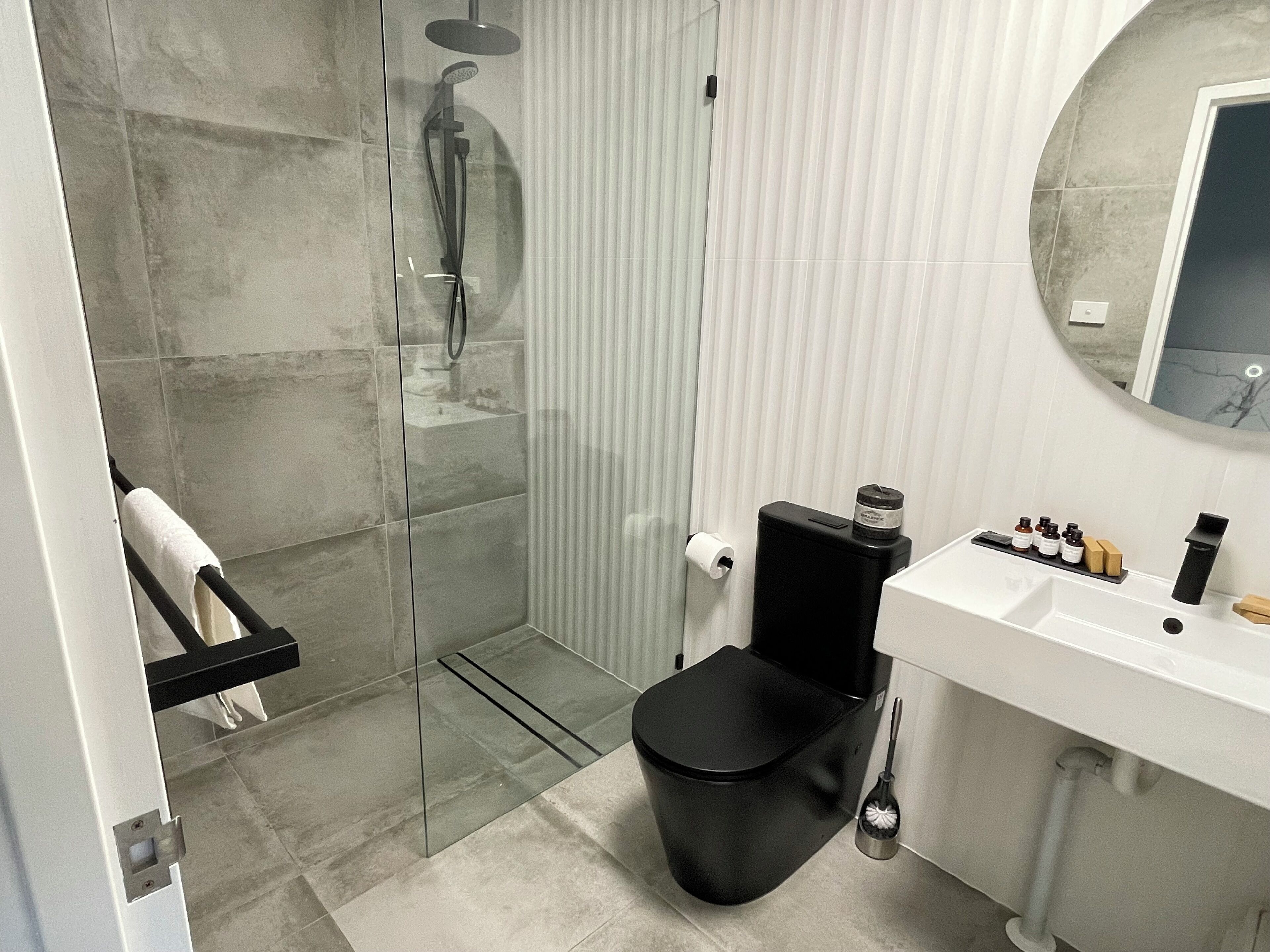 Une salle de bain moderne avec une douche à parois de verre, des toilettes noires, un lavabo blanc avec des articles de toilette et un miroir.
