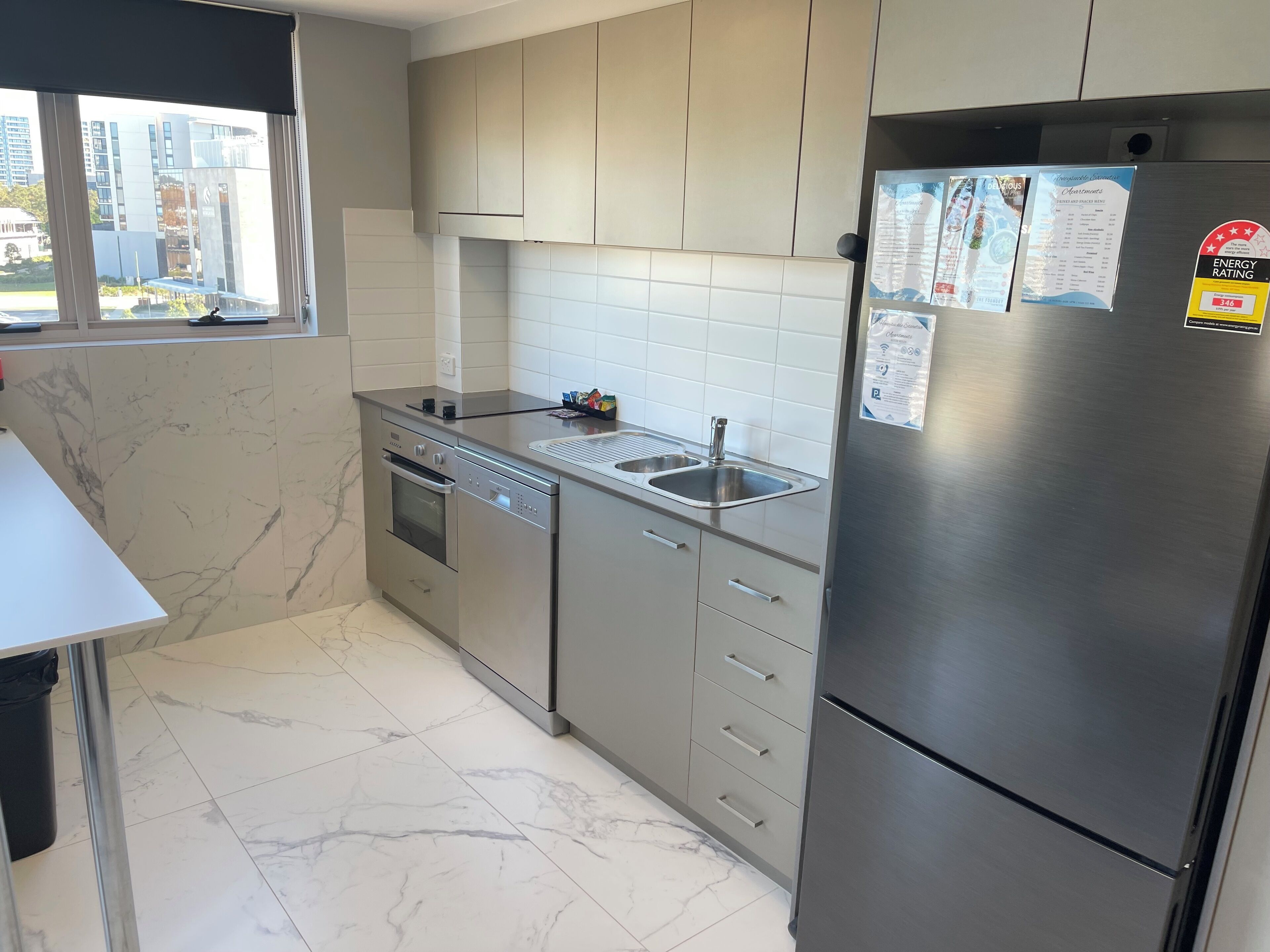 Una cocina moderna con refrigerador de acero inoxidable, horno empotrado y lavavajillas. Se aprecia una ventana con vista a la ciudad.