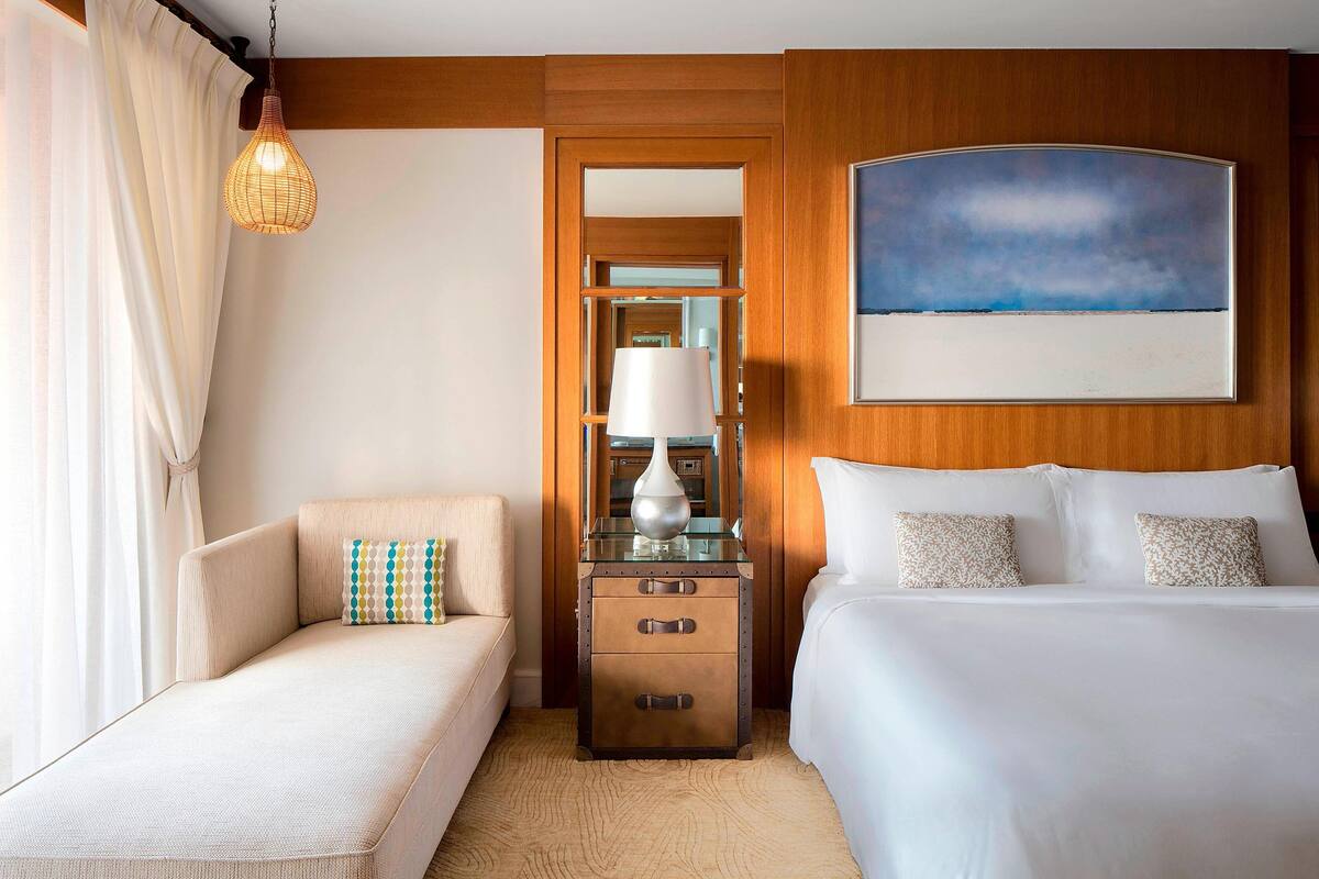 A hotel room with a bed, a sofa, a nightstand with drawers, a lamp, and a painting on the wall.