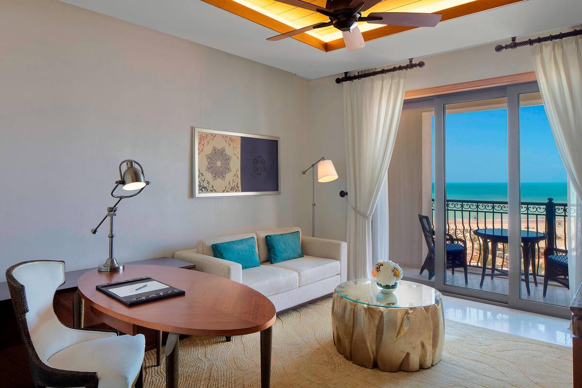 A hotel room with a balcony, a sofa, a dining table, and a ceiling fan.