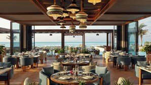 Un restaurante con mesas y sillas redondas, ventanas amplias y lámparas colgantes.