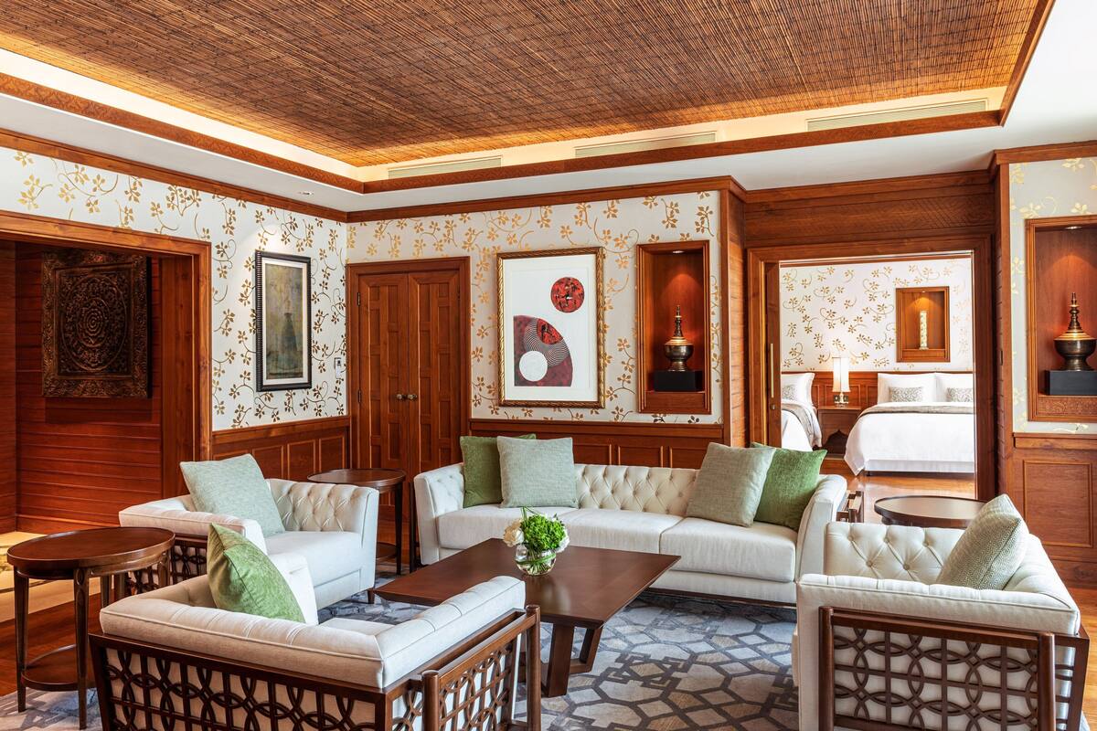 A hotel lobby with a large sofa, armchairs, a coffee table, and a dining table. There are framed artworks on the walls and a bed in the background.