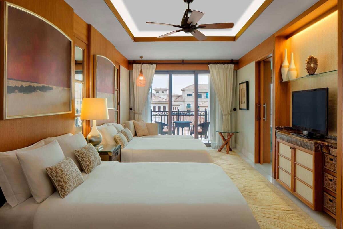 A hotel room with two beds, a TV, a ceiling fan, and a balcony with a table and chairs.