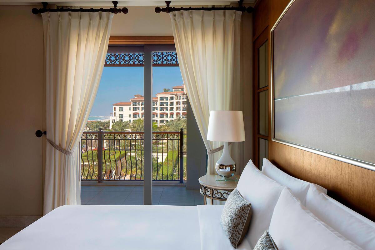 A hotel room with a balcony, a bed with white linens, a lamp, and a view of a building across the balcony.