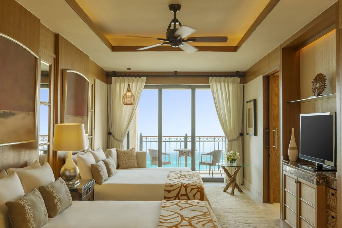 A hotel room with a large bed, a ceiling fan, a TV, a balcony with a view of the sea, and a small table with chairs.