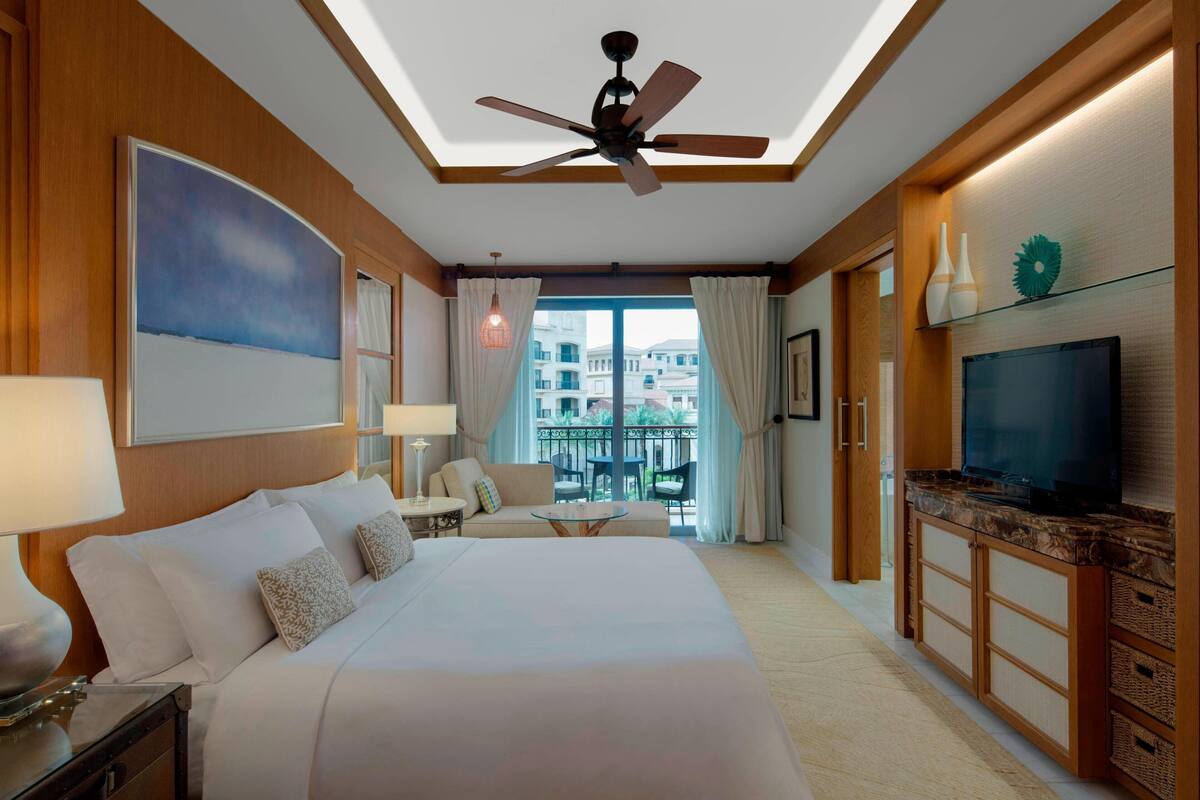 A hotel room with a large bed, a television, a ceiling fan, a balcony with a view of buildings, and a dining area with a table and chairs.