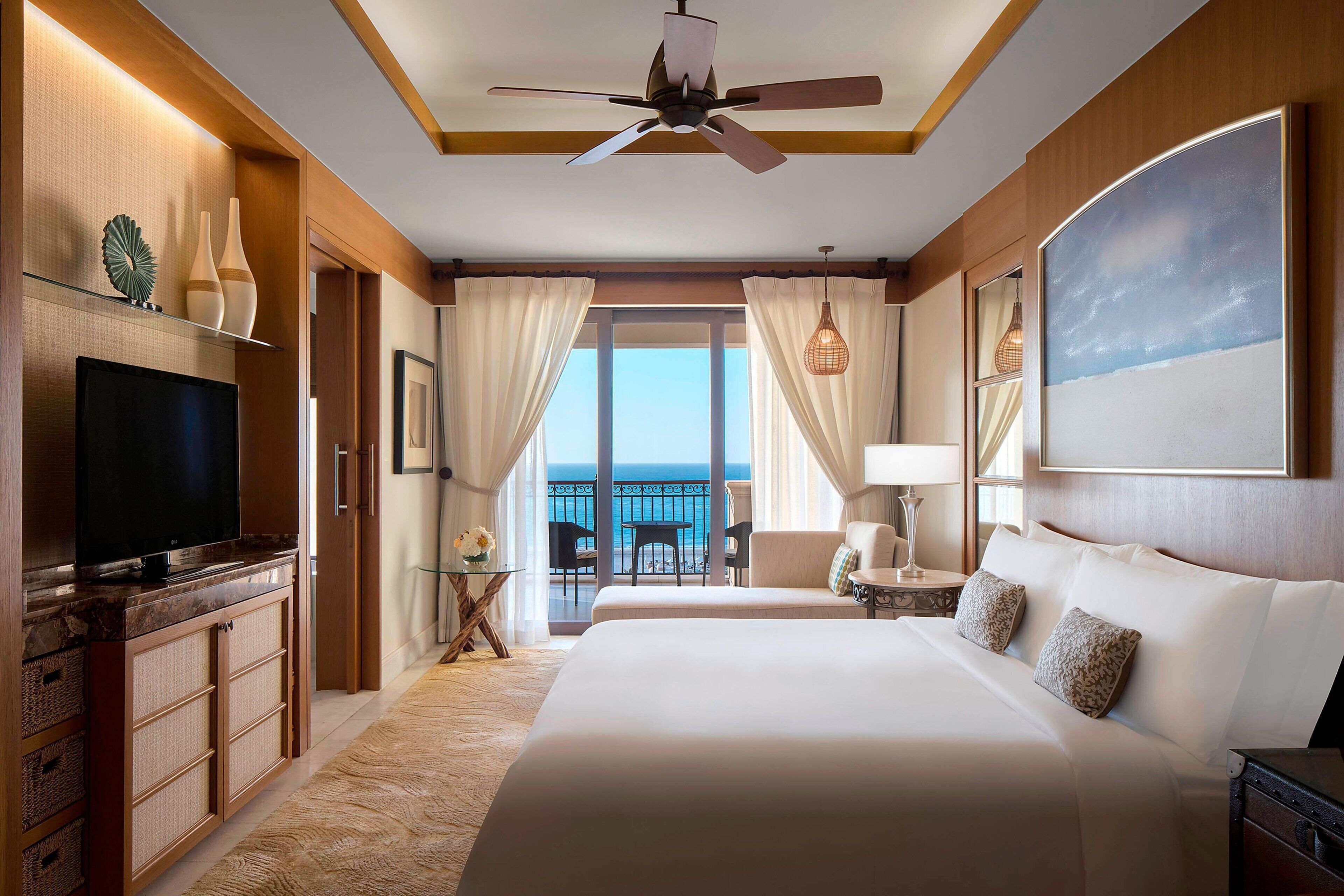 A hotel room with a bed, a television, a ceiling fan, a balcony with a view of the sea, and a painting on the wall.