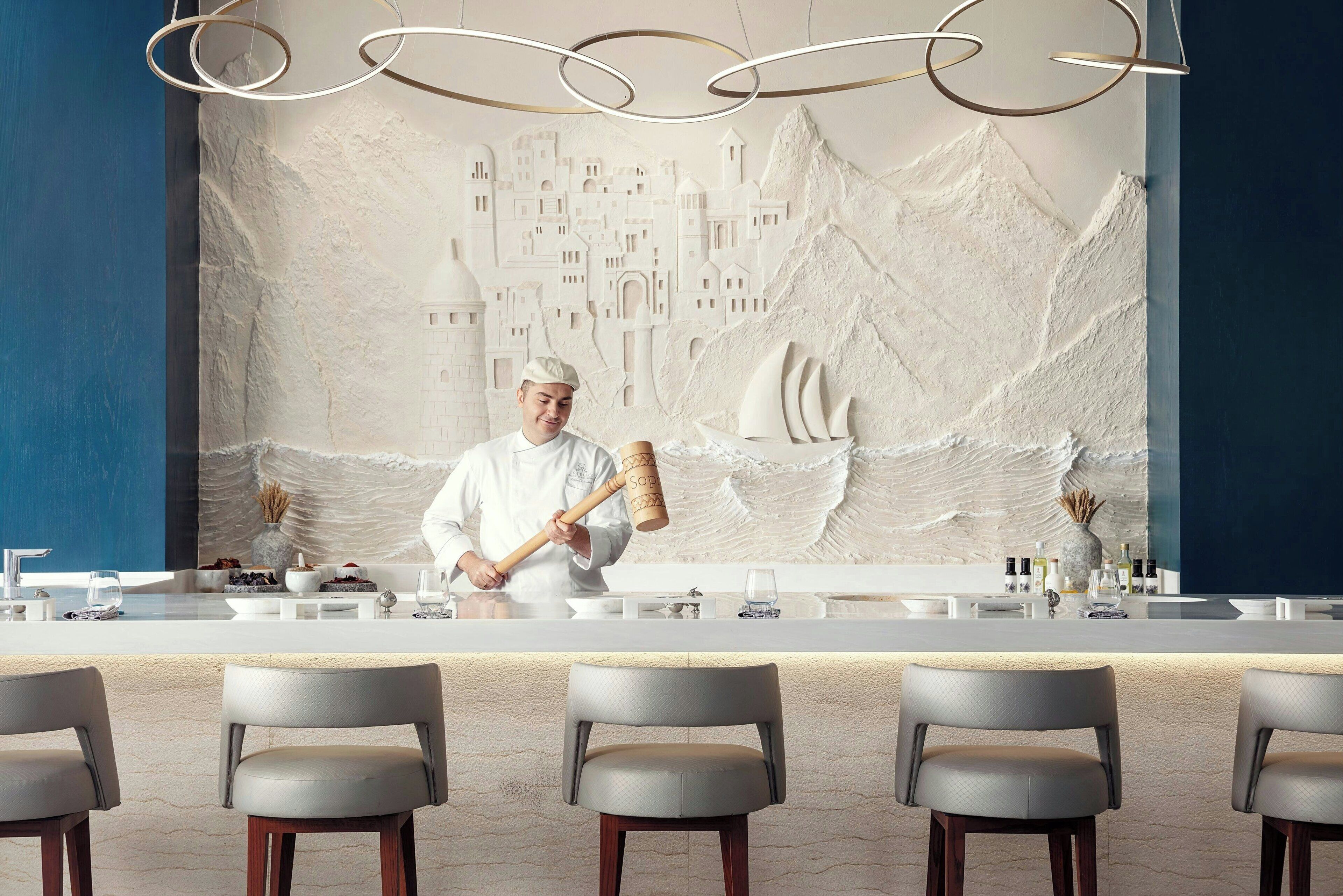 A chef at a counter with a cityscape mural behind him, bar stools in front, and a modern light fixture above.