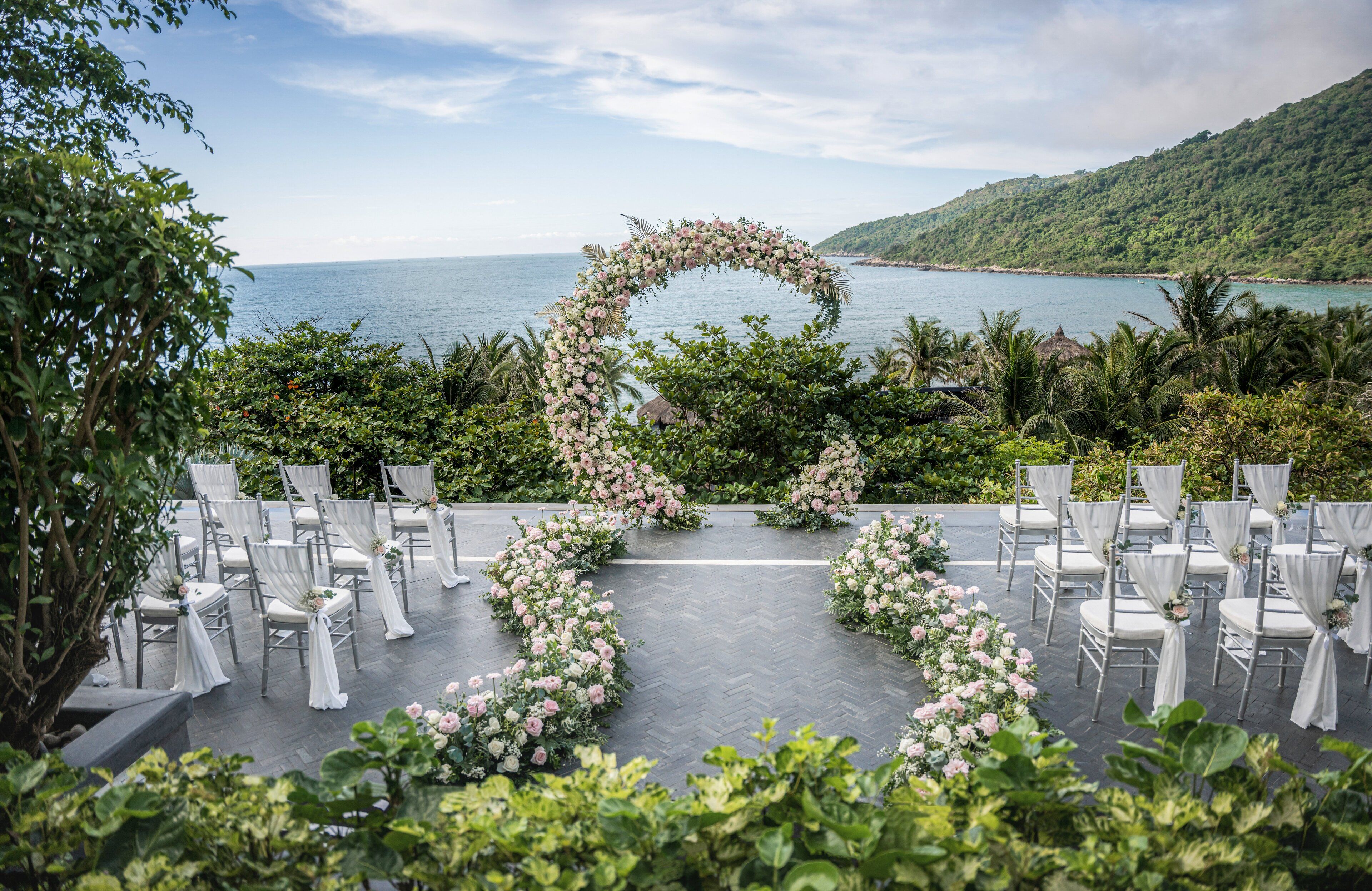 Un espacio para bodas al aire libre con un arco floral en forma de corazón, filas de sillas blancas y vistas al mar.