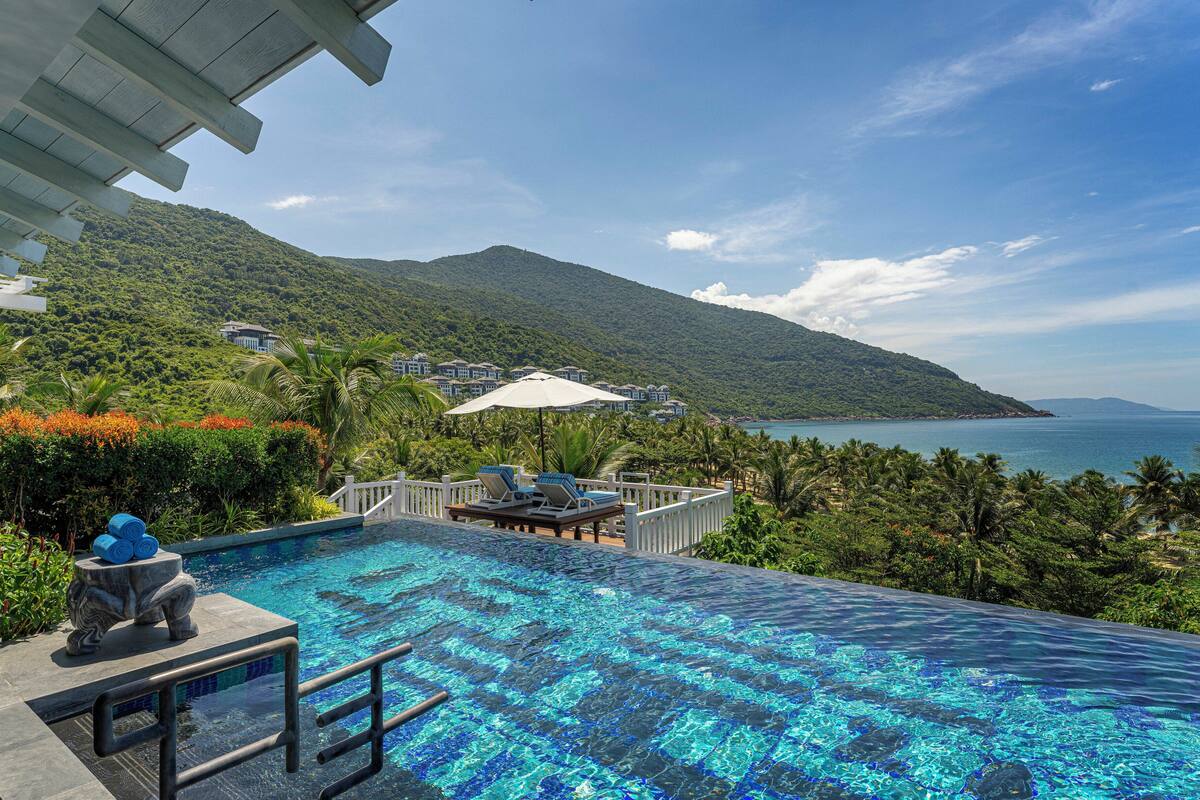 A infinity pool with a view of a mountain and the sea, surrounded by lush greenery and a white fence.
