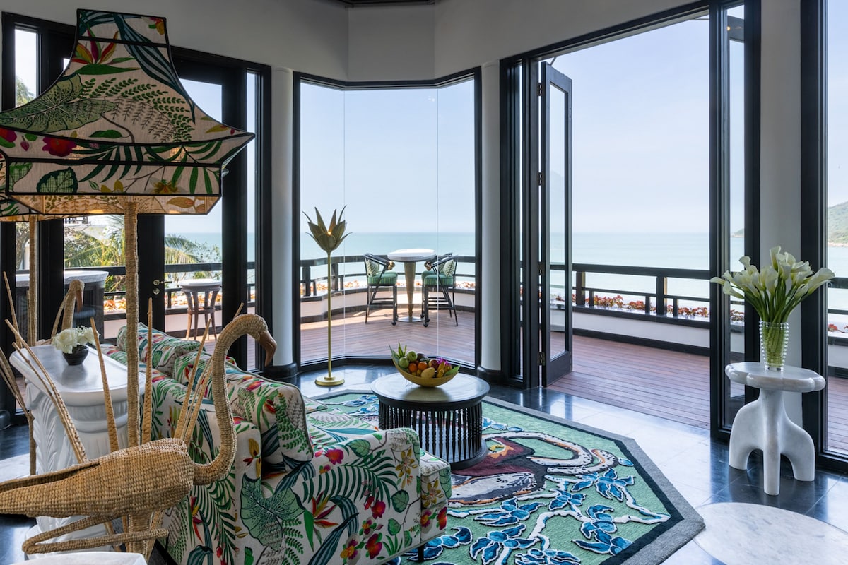 A spacious room with a large window overlooking the sea, a woven chair, a round table with a fruit bowl, and a decorative rug.