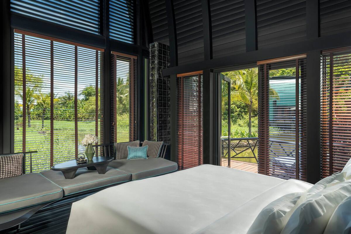 A modern bedroom with a large bed, a seating area with a table, and large windows offering a view of a garden.