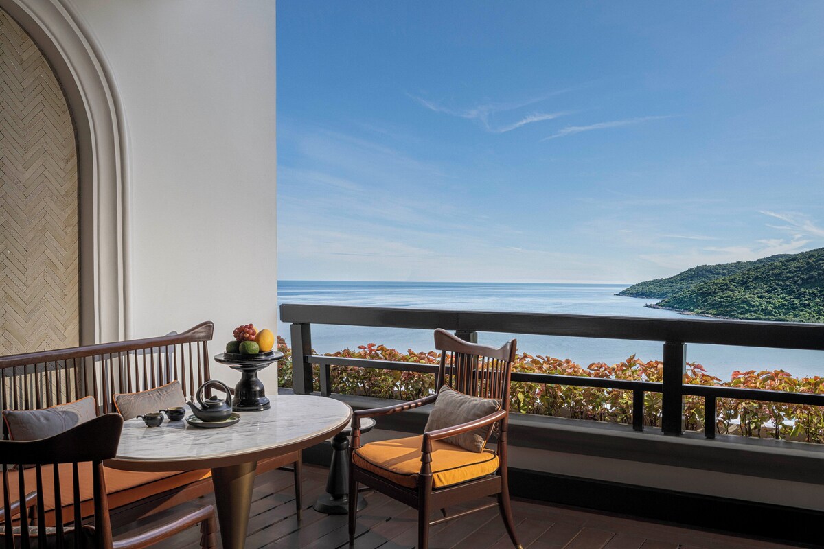 A balcony with a view of the sea, a round table with chairs, and a fruit bowl.