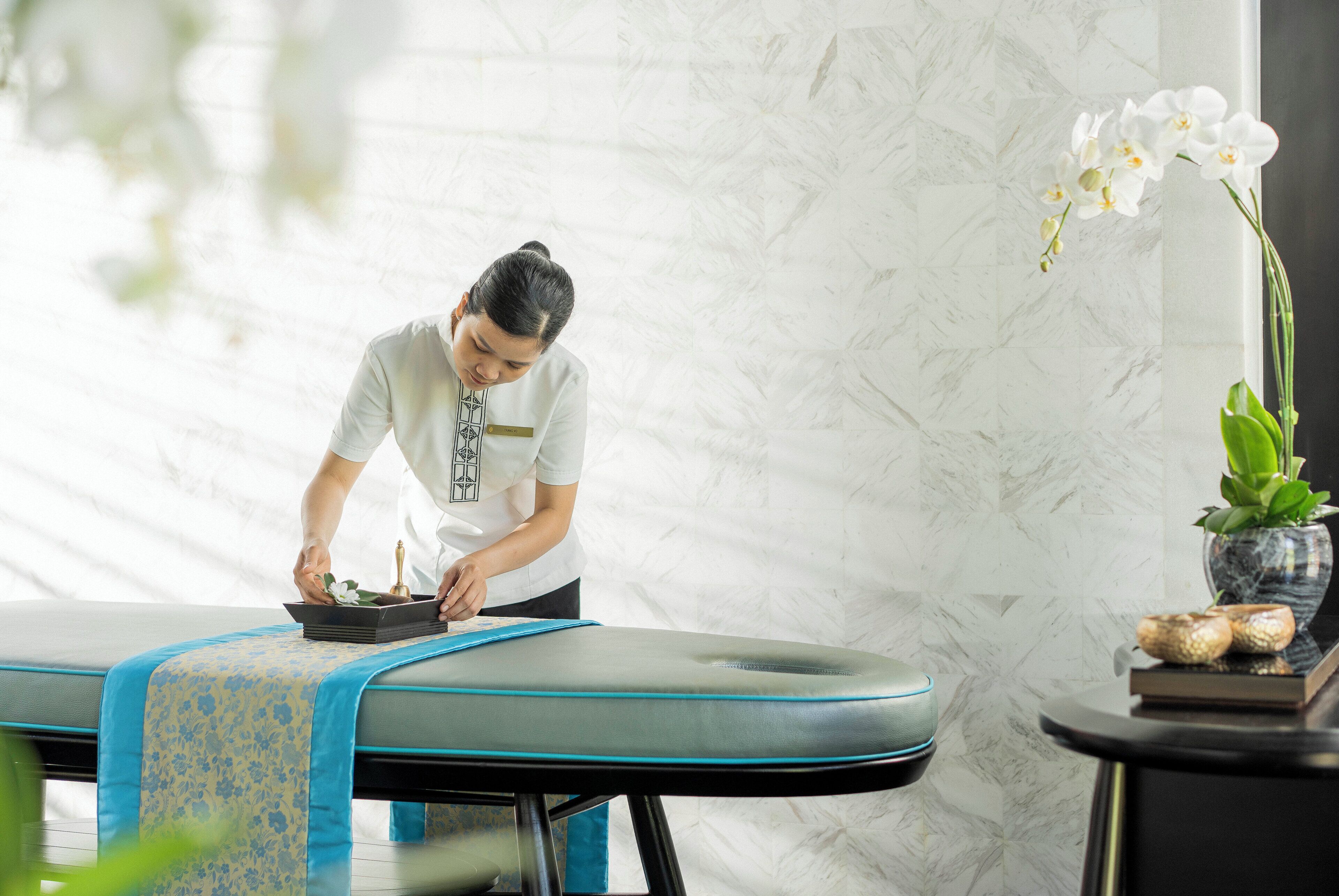A massage table with a blue and yellow patterned cloth, a vase with white flowers, and a small plant on a side table.