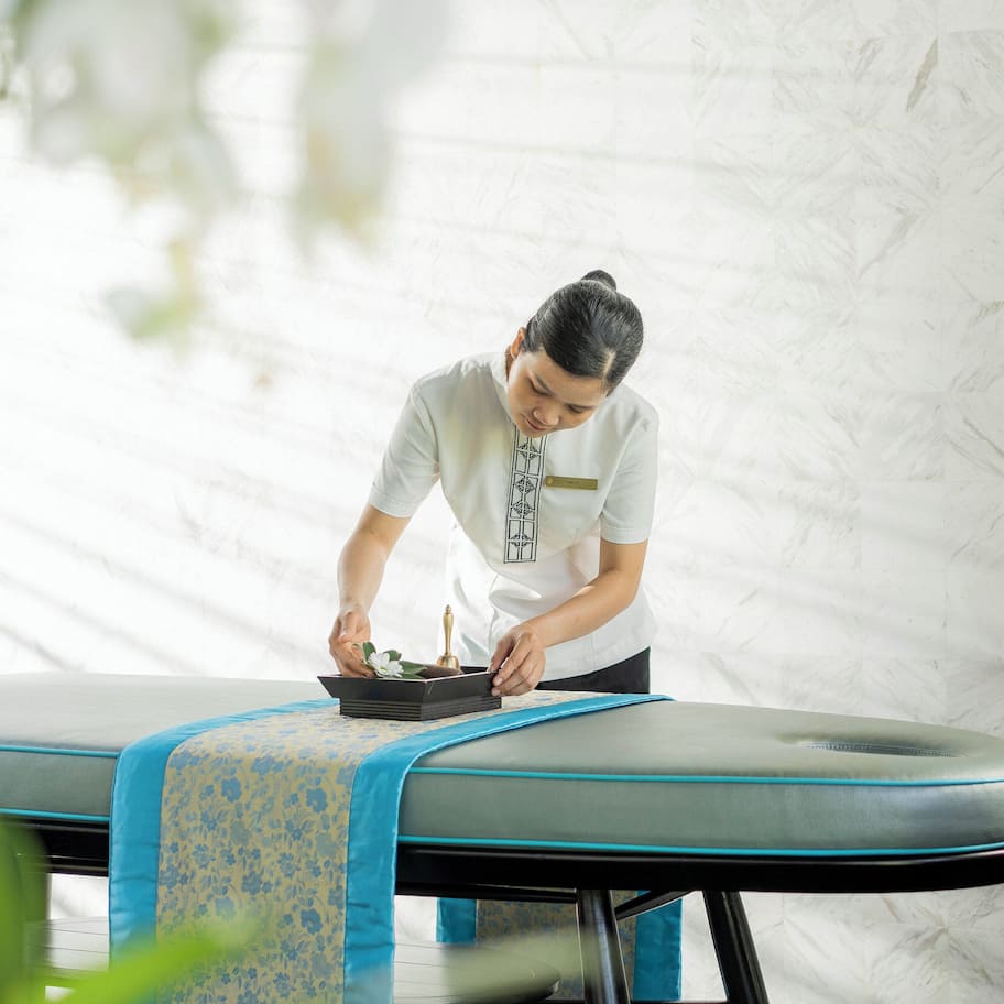 A massage table with a blue and yellow patterned cloth, a vase with white flowers, and a small plant on a side table.