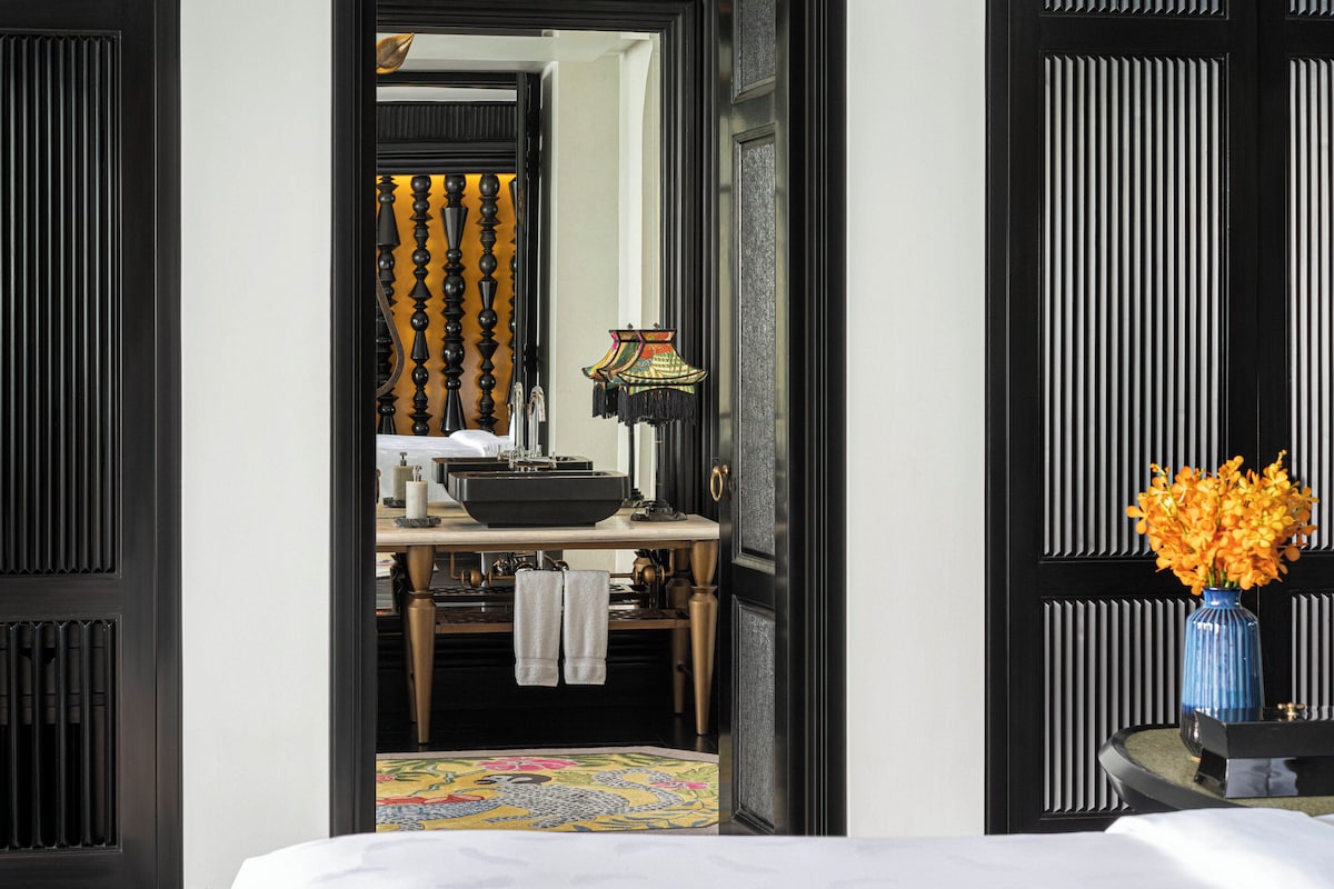 A hotel room with a bed, a bathroom with a sink and mirror, and a vase with flowers on a side table.