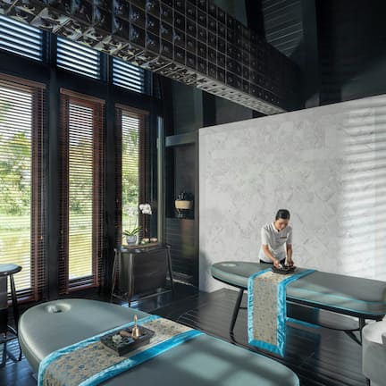 A modern spa room with a massage table, a small table with a plant, and a large window with blinds.