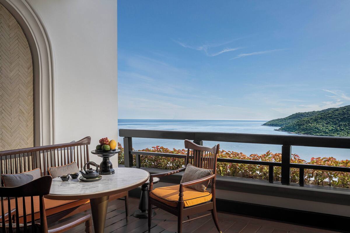 A balcony with a view of the sea, a round table with chairs, and a fruit bowl.