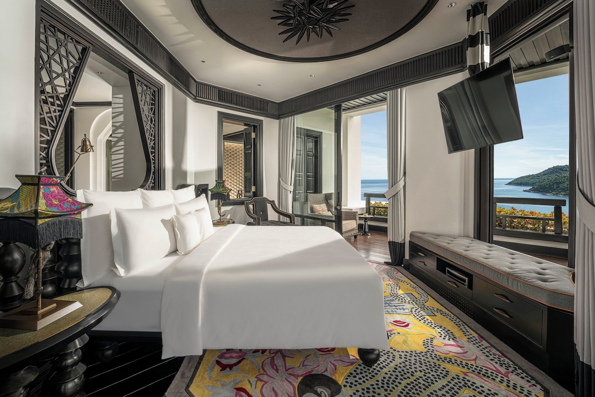A spacious bedroom with a large bed, a TV, and a balcony with a view of the sea.