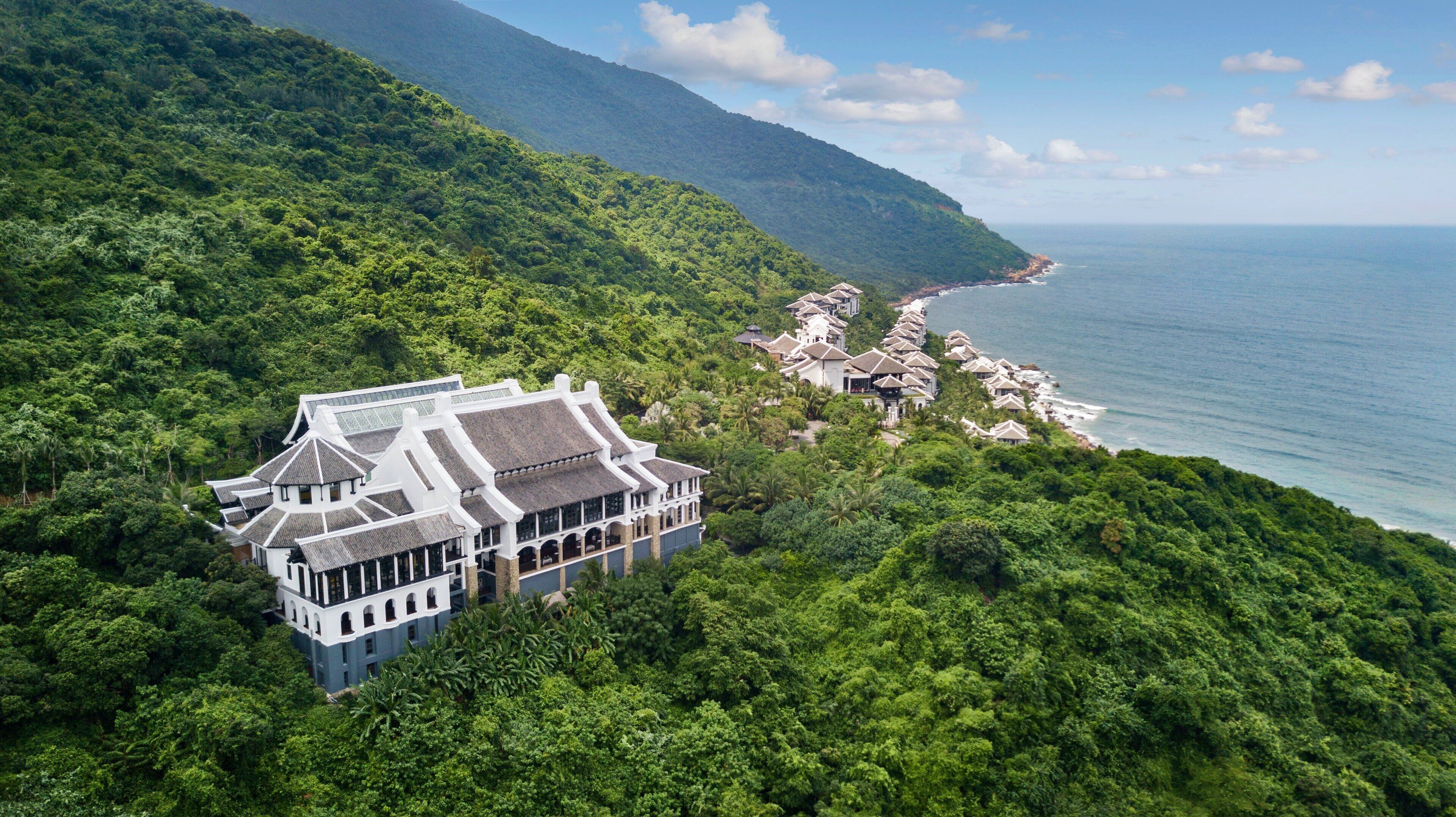A traditional building with multiple levels and a large balcony, surrounded by dense greenery and overlooking a coastline.