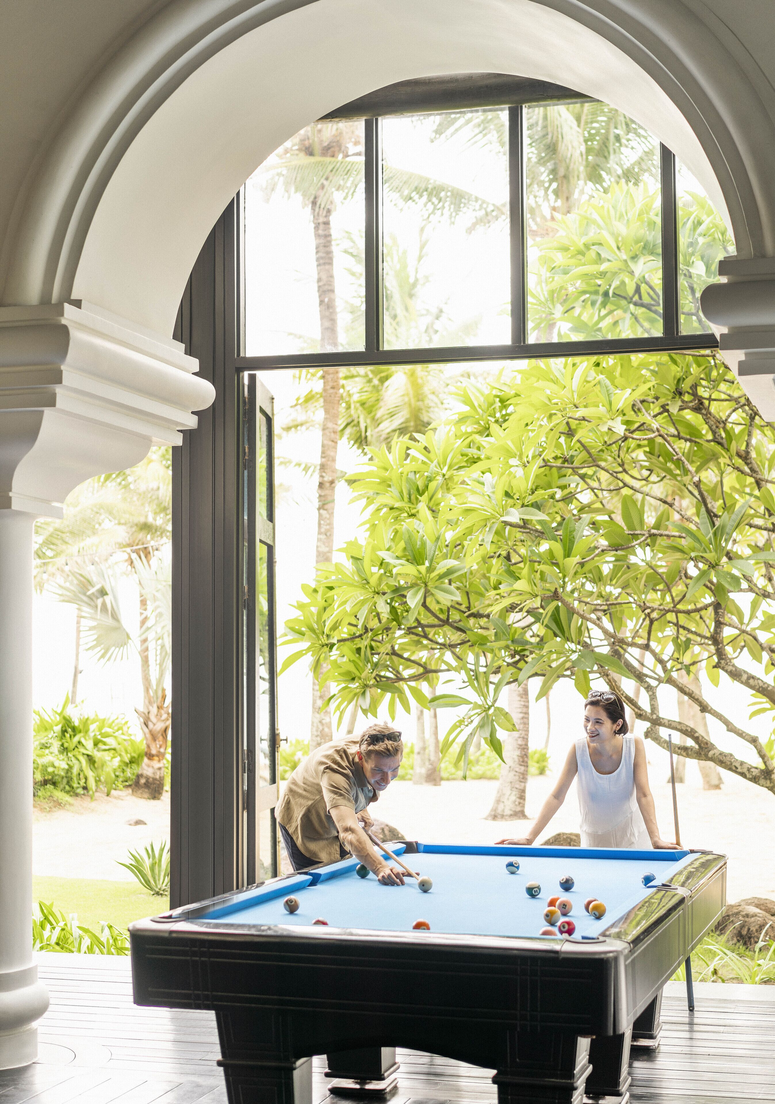 A pool table in a covered outdoor area with a view of tropical plants and palm trees.