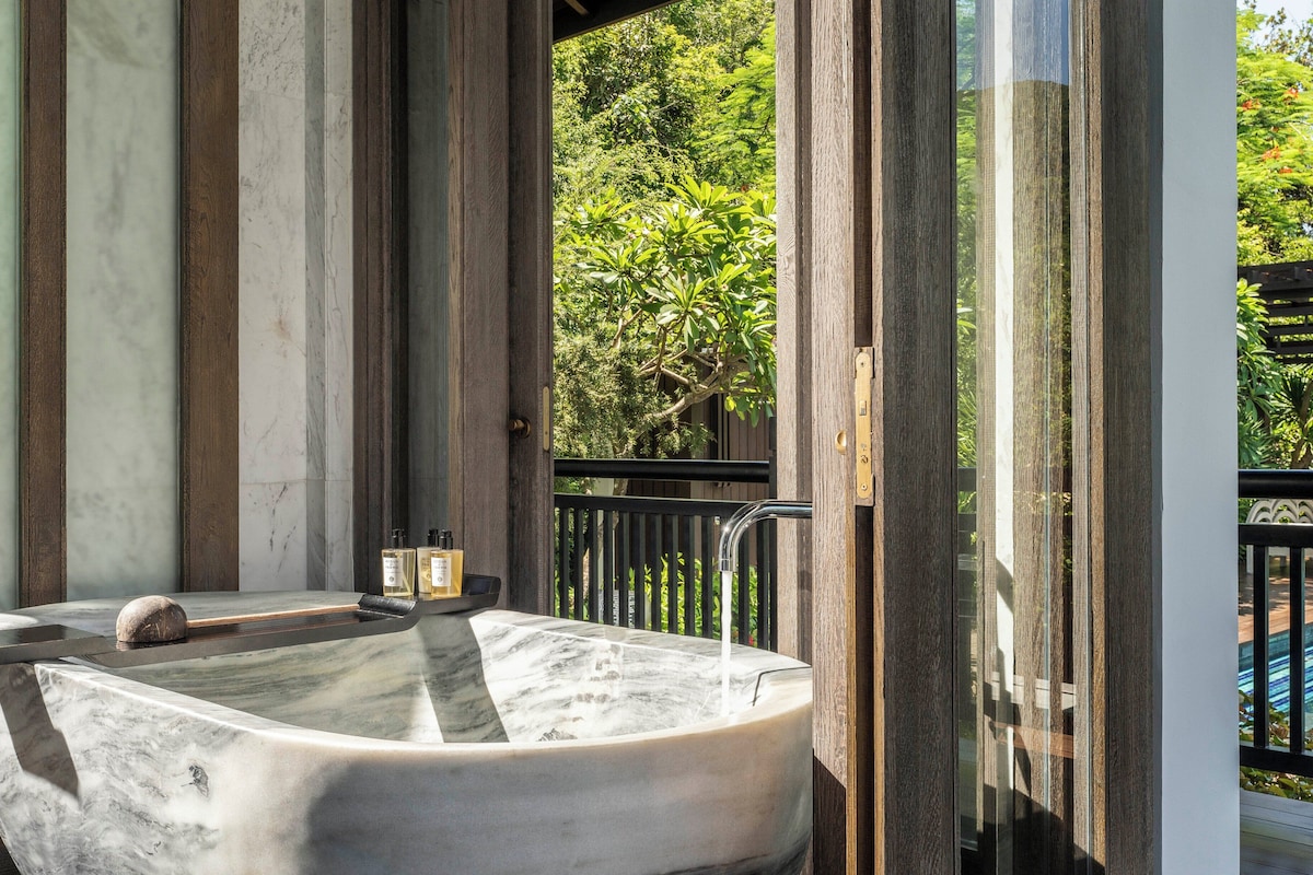 A spa area with a large marble bathtub, wooden columns, and a view of lush greenery.