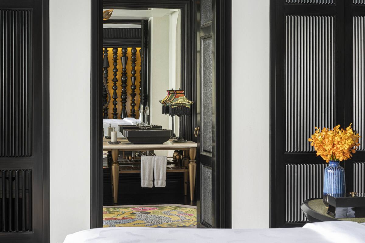 A hotel room with a bed, a bathroom with a sink and mirror, and a vase with flowers on a side table.