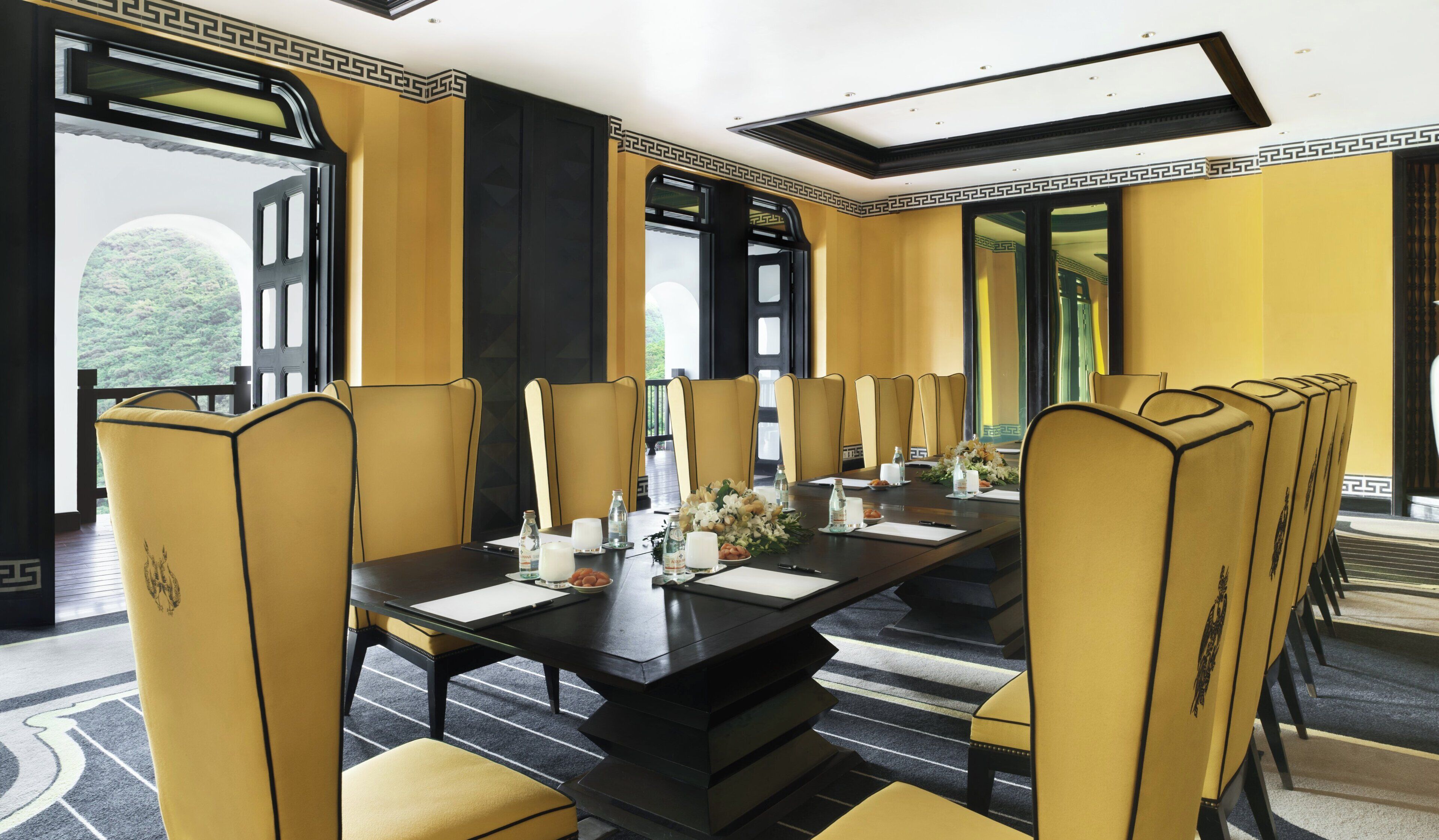 A formal dining room with a long table, multiple chairs, and a view of greenery outside.