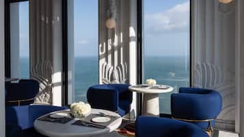 A modern dining area with blue upholstered chairs, white tables, and a view of the ocean through large windows.