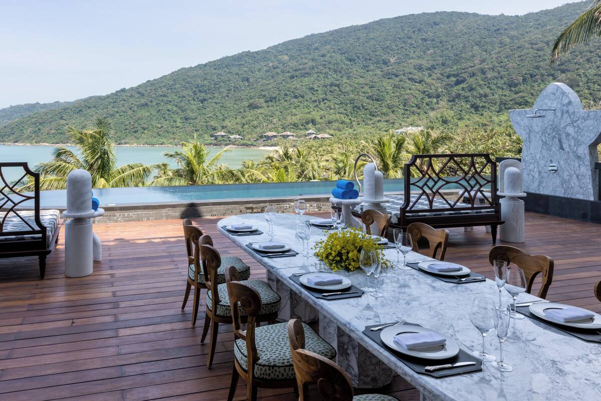 A dining area with a long table set for six, overlooking a pool and lush greenery.