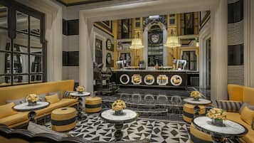 A hotel lobby with a geometric patterned floor, yellow sofas, round tables with flower arrangements, and a central reception area with a large mirror and decorative elements.