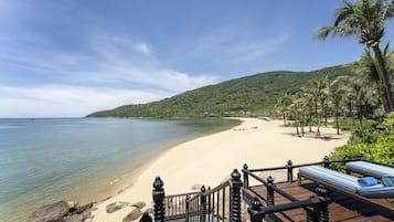 Eine Holzterrasse mit Geländer, die auf einen Strand mit klarem Wasser und einem Berg im Hintergrund blickt.