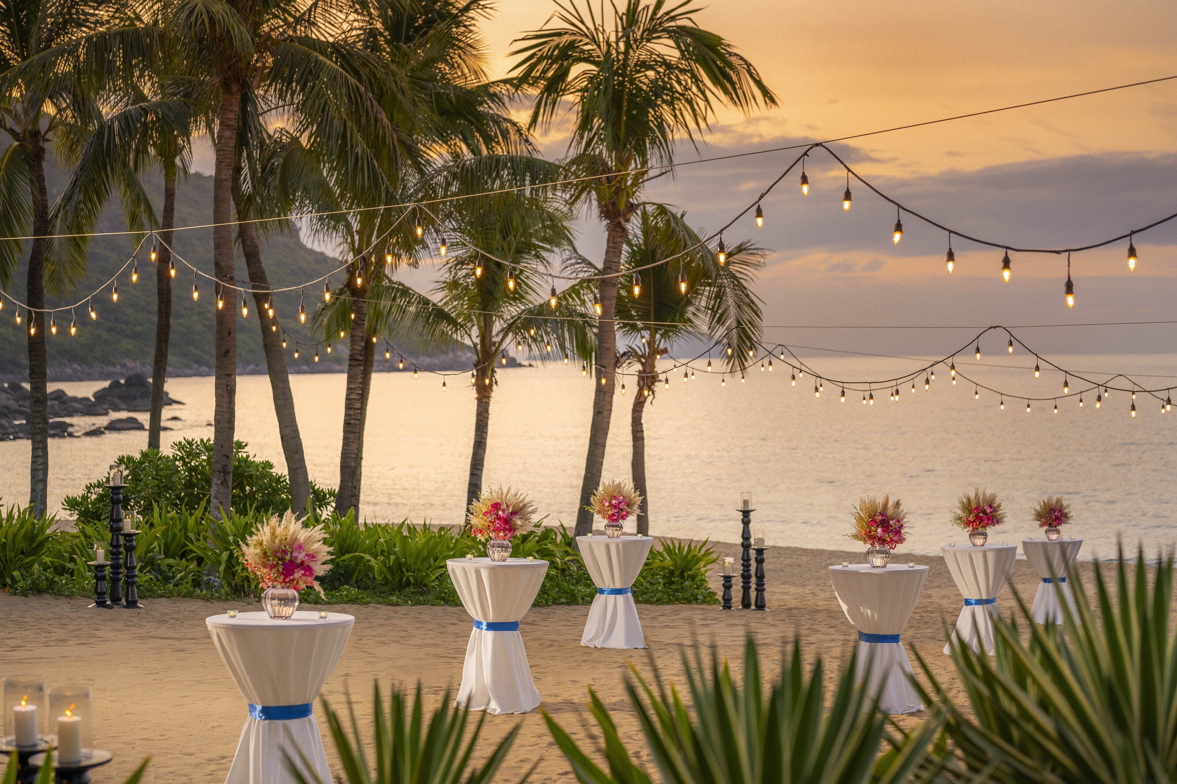 Un entorno frente al mar con luces colgantes, palmeras y mesas con centros de flores.