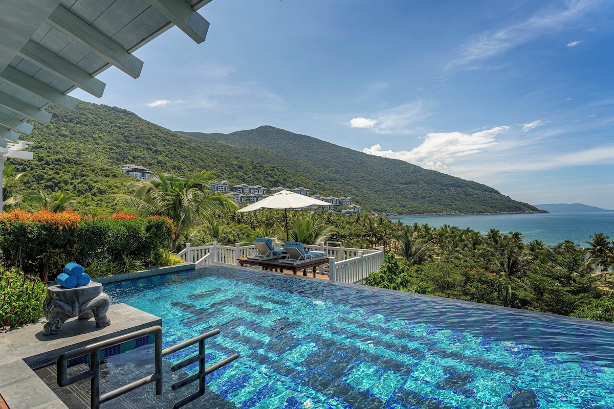 A infinity pool with a view of a mountain and the sea, surrounded by lush greenery and a white fence.